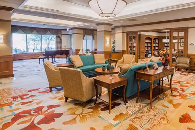 A spacious and well-lit living room area in a senior living facility featuring comfortable seating including a teal sofa and beige armchairs arranged around wooden tables. The room has large windows with a view of trees outside, a grand piano near the windows, and a bookshelf filled with books. The floor is covered with a colorful patterned carpet, and the walls have wooden paneling and decorative lighting.