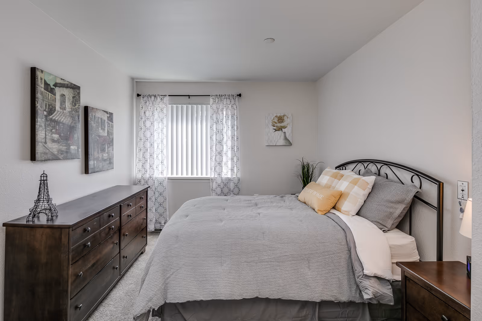 Neatly furnished bedroom with a bed made in gray linens, wooden dressers, wall art and a window with patterned curtains.