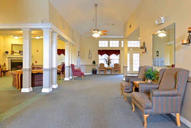 A spacious and well-lit common area in a senior living facility with high ceilings, ceiling fans, and large windows. The room features comfortable striped armchairs with a small table and plant, a large mirror on the wall, and a seating area with couches and a fireplace visible in the background. There are also dining tables and chairs near the windows with red valances.
