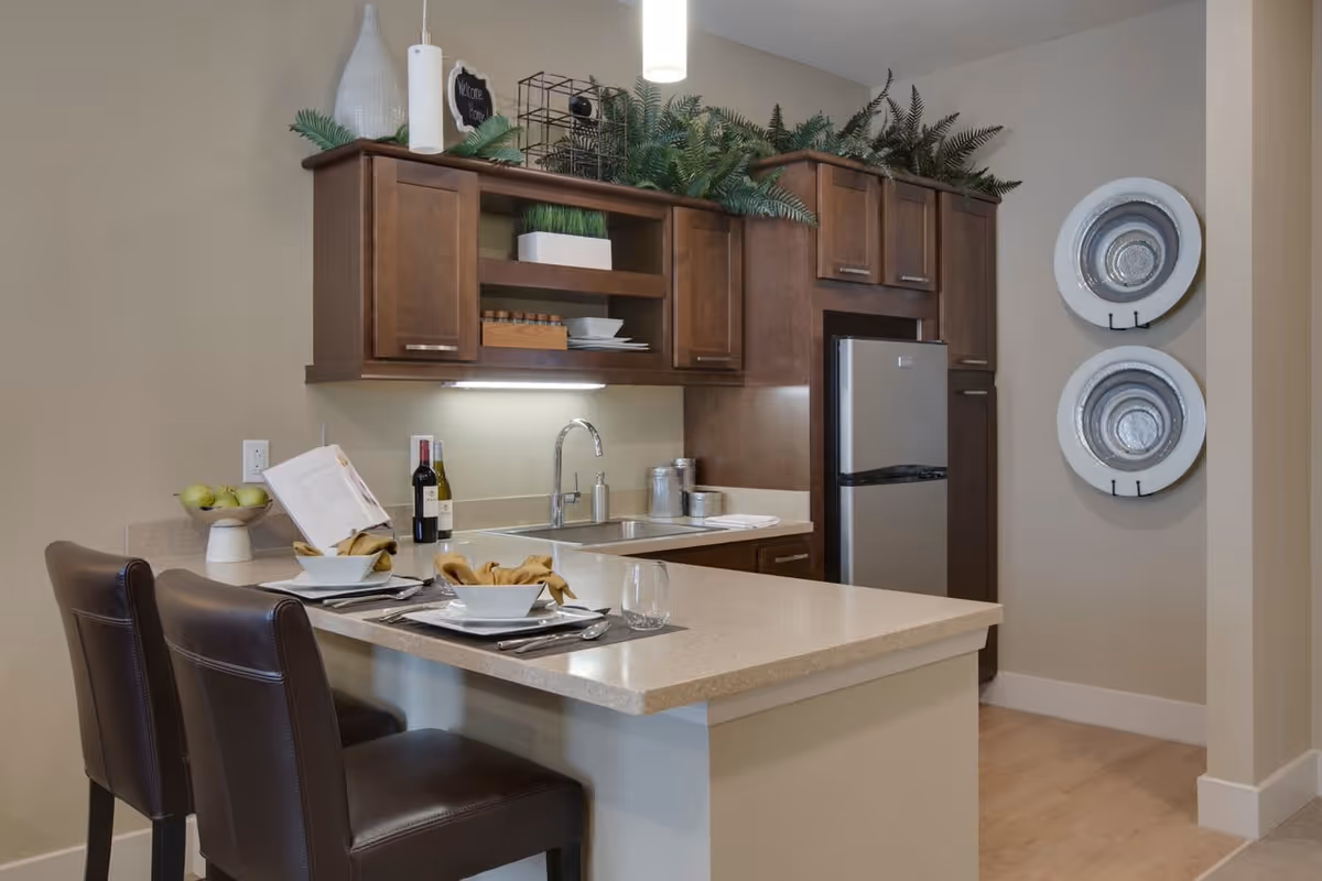 Modern kitchen area with a beige countertop island featuring two brown leather chairs set with white bowls, napkins, and glasses. The kitchen has dark wooden cabinets, a stainless steel refrigerator, a sink with a faucet, and decorative plants on top of the cabinets. Two large decorative plates are mounted on the wall to the right.