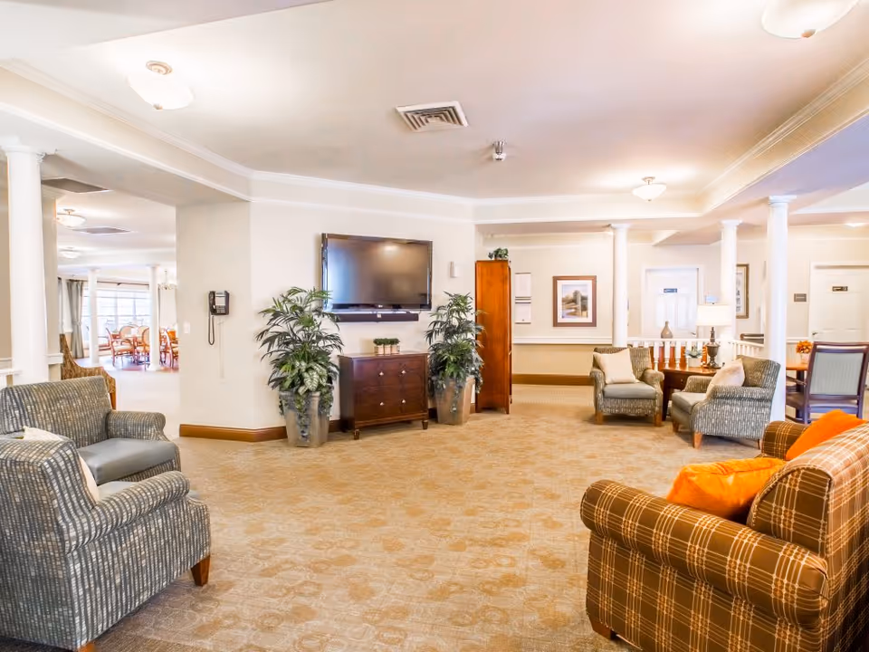 A spacious senior living facility common area with comfortable armchairs and sofas arranged around a flat-screen TV mounted on the wall. The room features beige carpeting, white walls, decorative plants, and several columns. In the background, there is a dining area with tables and chairs near large windows.