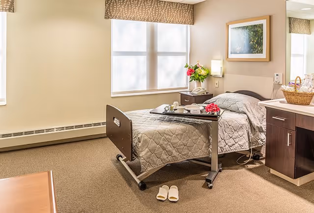 A neatly arranged senior living bedroom with a single hospital-style bed covered in a beige quilt. A tray with a cup, plate, and napkin is placed on an adjustable overbed table. There is a pair of white slippers on the carpeted floor near the bed. The room has a window with a patterned valance, a nightstand with a telephone and a vase of flowers, a framed picture on the wall, and a cabinet with a basket on top.