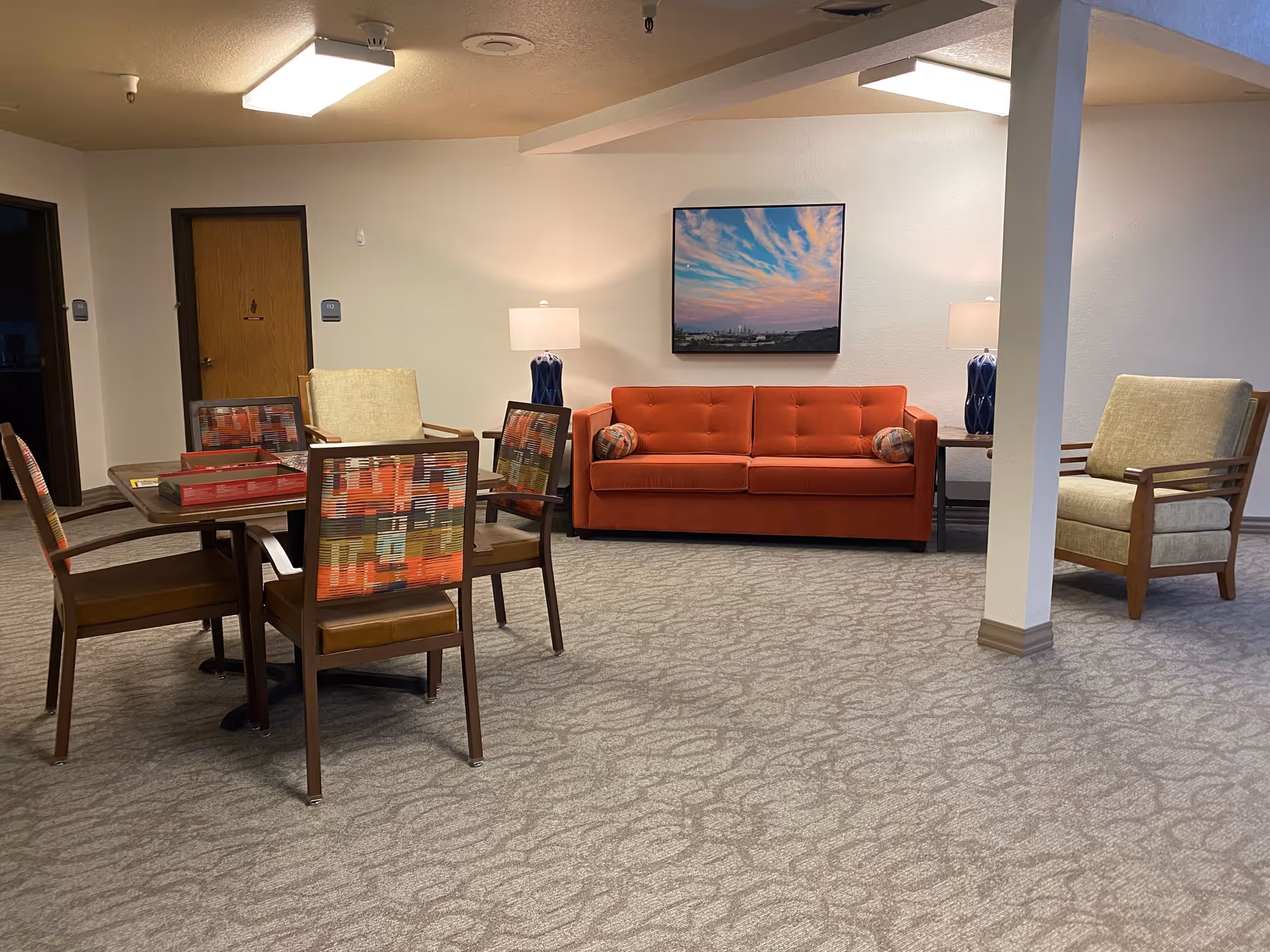 A senior living facility common area with a red sofa, two beige armchairs, a wooden table with four chairs featuring colorful patterned backs, two table lamps with blue bases, and a framed picture of a sunset sky on the wall.
