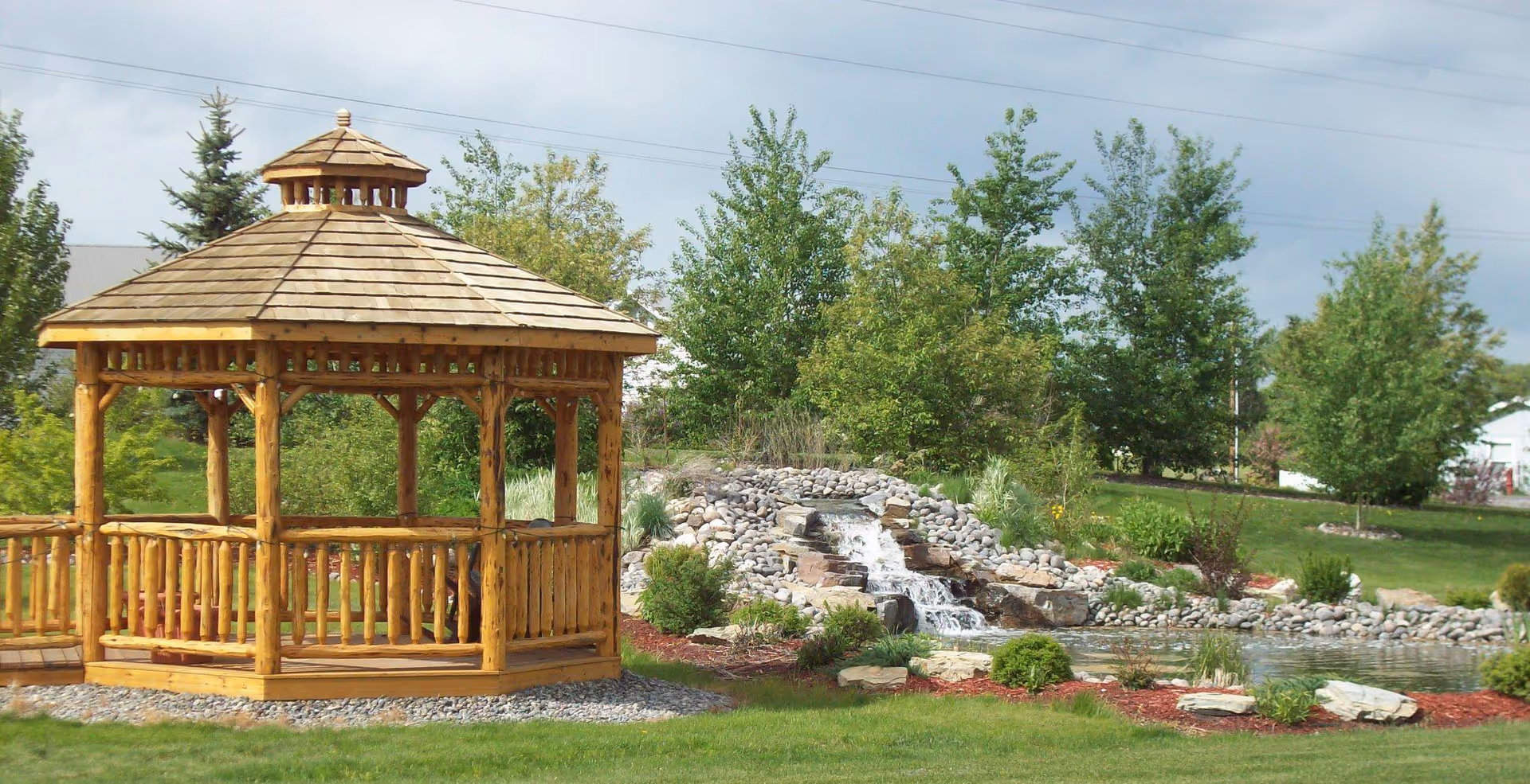Wooden gazebo next to a landscaped pond with a small rock waterfall and surrounding trees and lawn.