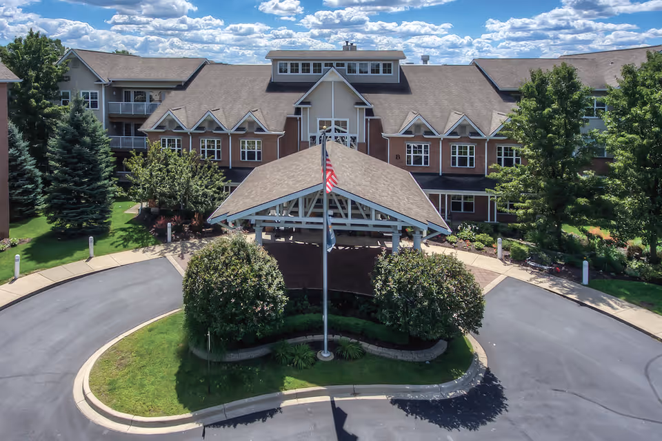 Front exterior view of Town Village Sterling Heights senior living facility with a covered entrance, American flag on a flagpole, surrounded by trees and landscaping under a partly cloudy sky.