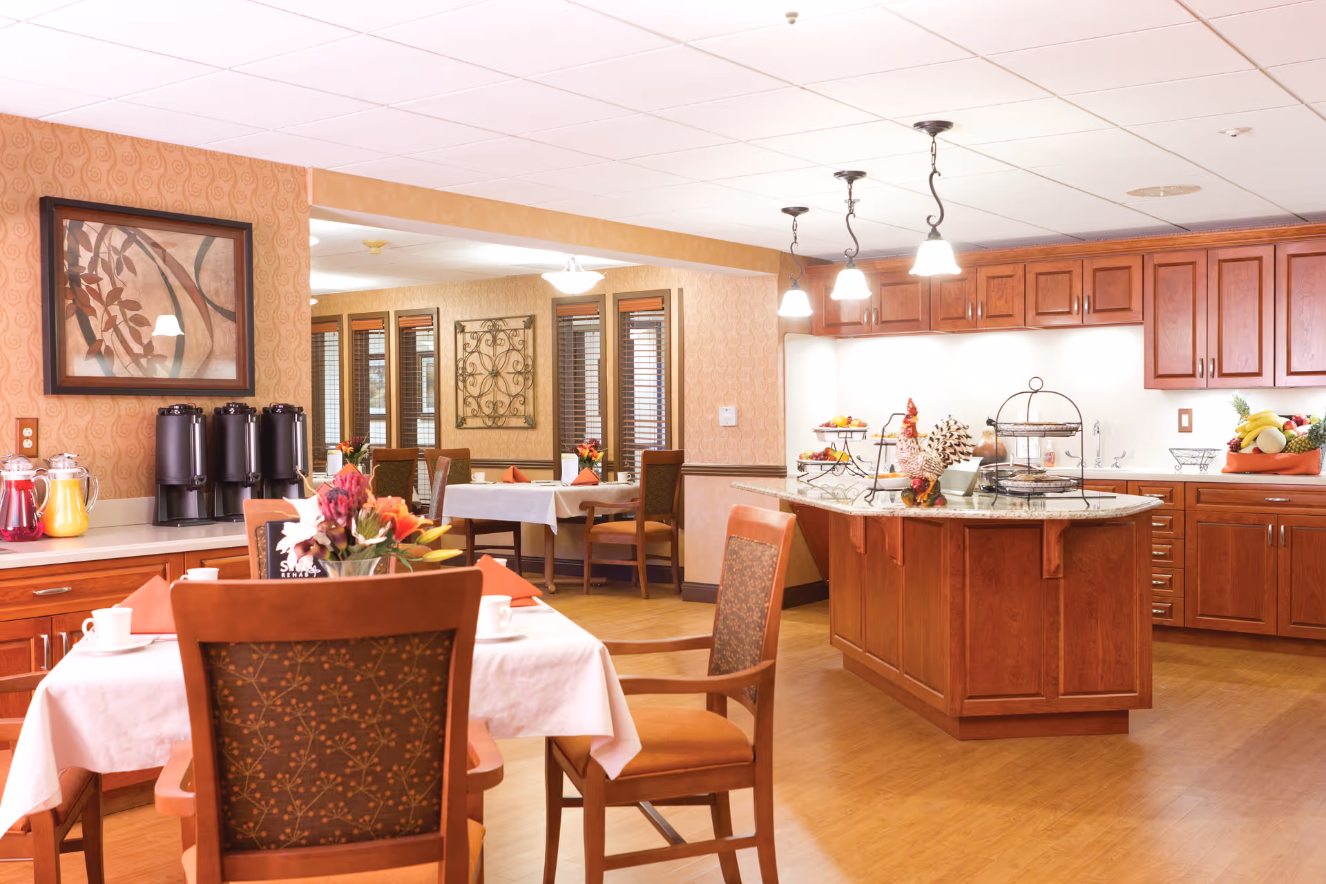 A warm and inviting dining area with wooden chairs and tables covered with white tablecloths and orange napkins. The room features a kitchen island with granite countertop, wooden cabinets, and hanging pendant lights. There are beverage dispensers and a floral centerpiece on one of the tables. The background shows additional dining tables and decorative wall art.