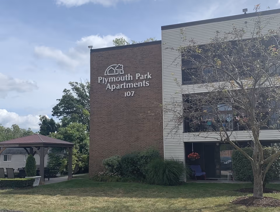 Exterior view of a brick apartment building with a sign reading "Plymouth Park Apartments 107", balconies, a lawn, and a nearby gazebo.