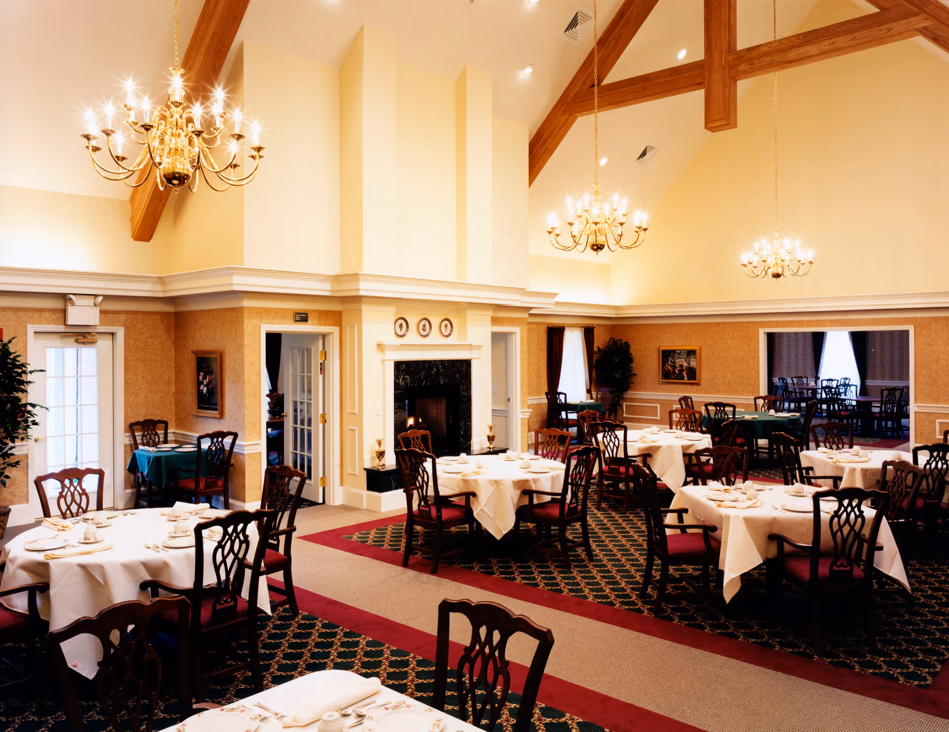 Well-lit formal dining room with round tables set with white tablecloths, chandeliers, and wooden chairs.