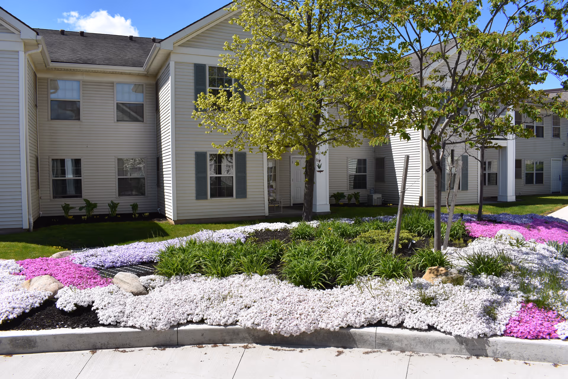Exterior view of a senior living facility building with beige siding and green shutters. In front of the building is a landscaped garden with blooming white, purple, and pink flowers, green shrubs, and several young trees. The sky is blue with a few clouds.