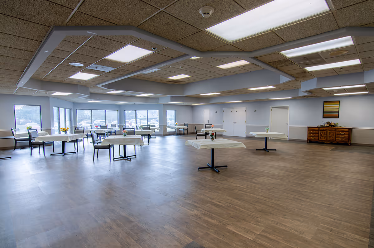 A spacious dining room with several tables covered with white tablecloths and small flower arrangements. The room has large windows letting in natural light, wooden flooring, and a ceiling with recessed lighting. There is a wooden sideboard with a flower arrangement and a framed picture on the wall.