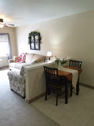 A cozy living area with a beige sofa and a patterned armchair separated by a half wall from a small dining area with a wooden table and two chairs. The table is decorated with a white runner and a small floral arrangement. A lamp sits on the half wall, and a window with blinds allows natural light into the room. A decorative wall hanging with greenery is above the sofa.