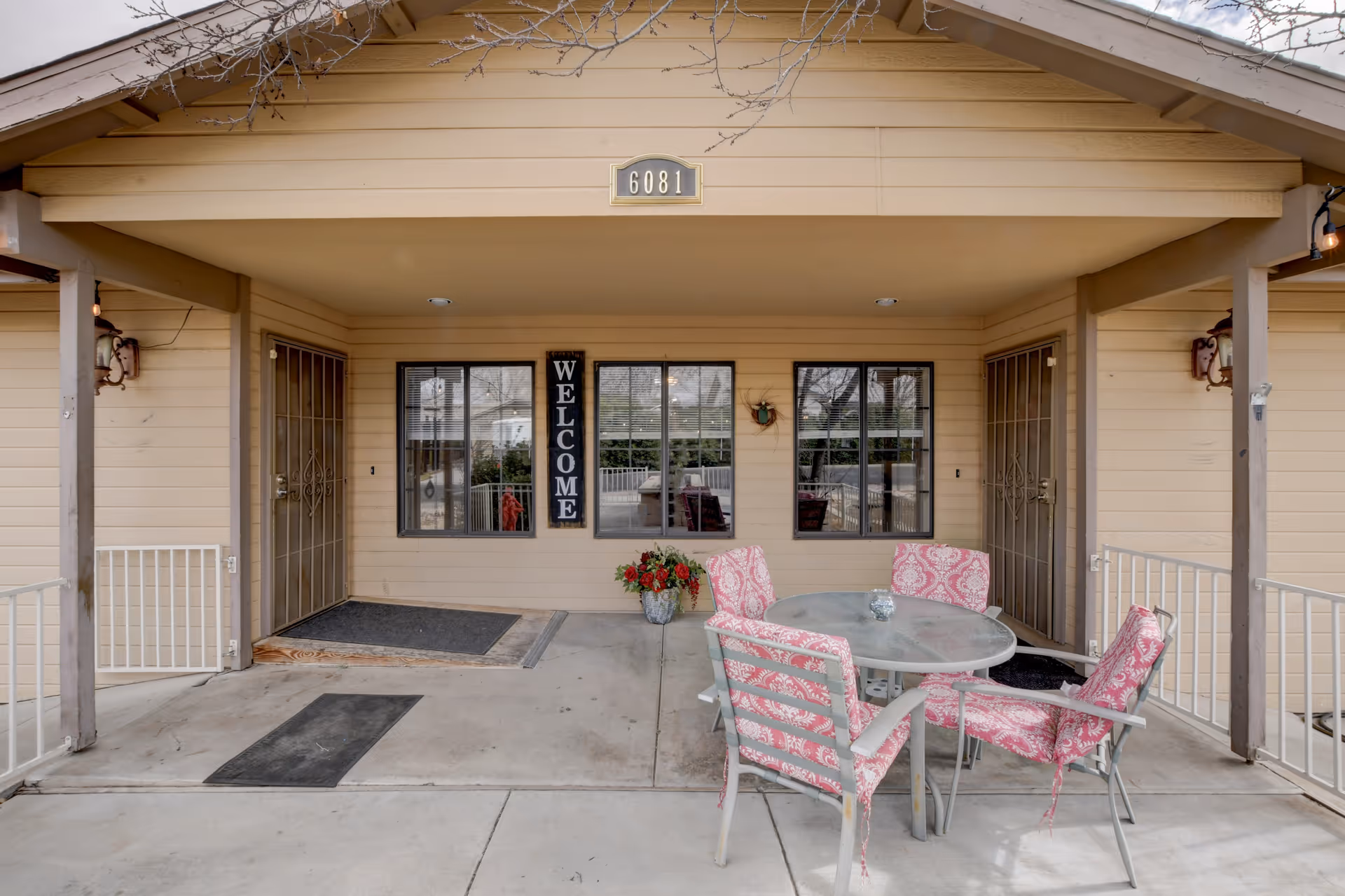 Covered porch area of a building with beige siding, featuring two doors with metal security screens on either side. Between the doors are three windows and a vertical sign that reads 'WELCOME'. In front of the windows is a round glass table with four chairs that have pink patterned cushions. A small flower pot with red flowers is placed near the windows. The building number '6081' is displayed above the porch.