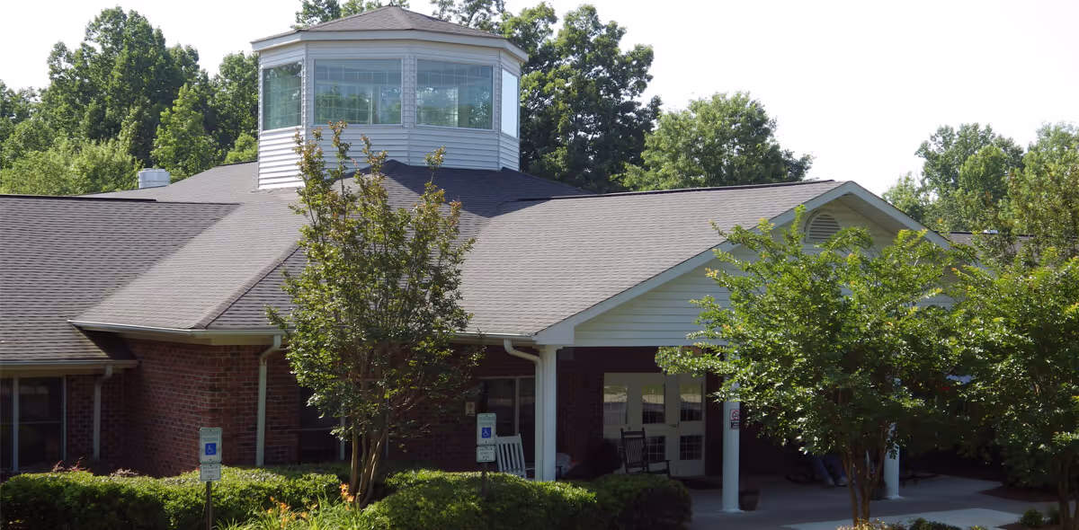 Exterior view of a single-story brick building with a gray shingled roof and a white cupola on top. The entrance is covered by a white portico supported by columns, with two rocking chairs visible near the door. Trees and shrubs surround the building, and there are two handicap parking signs near the entrance.