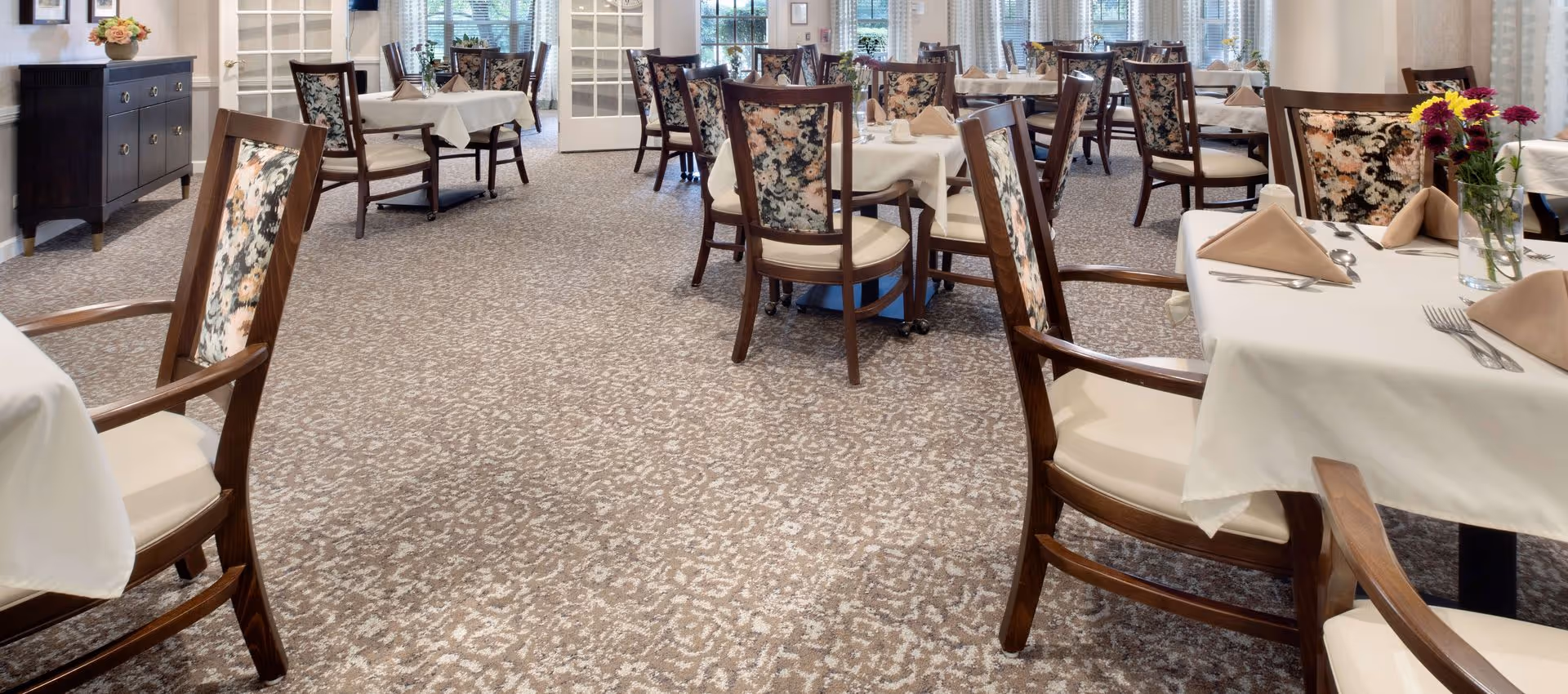 A dining room in a senior living facility with multiple tables covered in white tablecloths, each set with beige napkins, silverware, and small flower arrangements. The chairs have wooden frames with floral patterned upholstery on the backrests. The room has large windows with curtains allowing natural light to enter, and a dark wooden sideboard with a flower arrangement on top is visible on the left side.