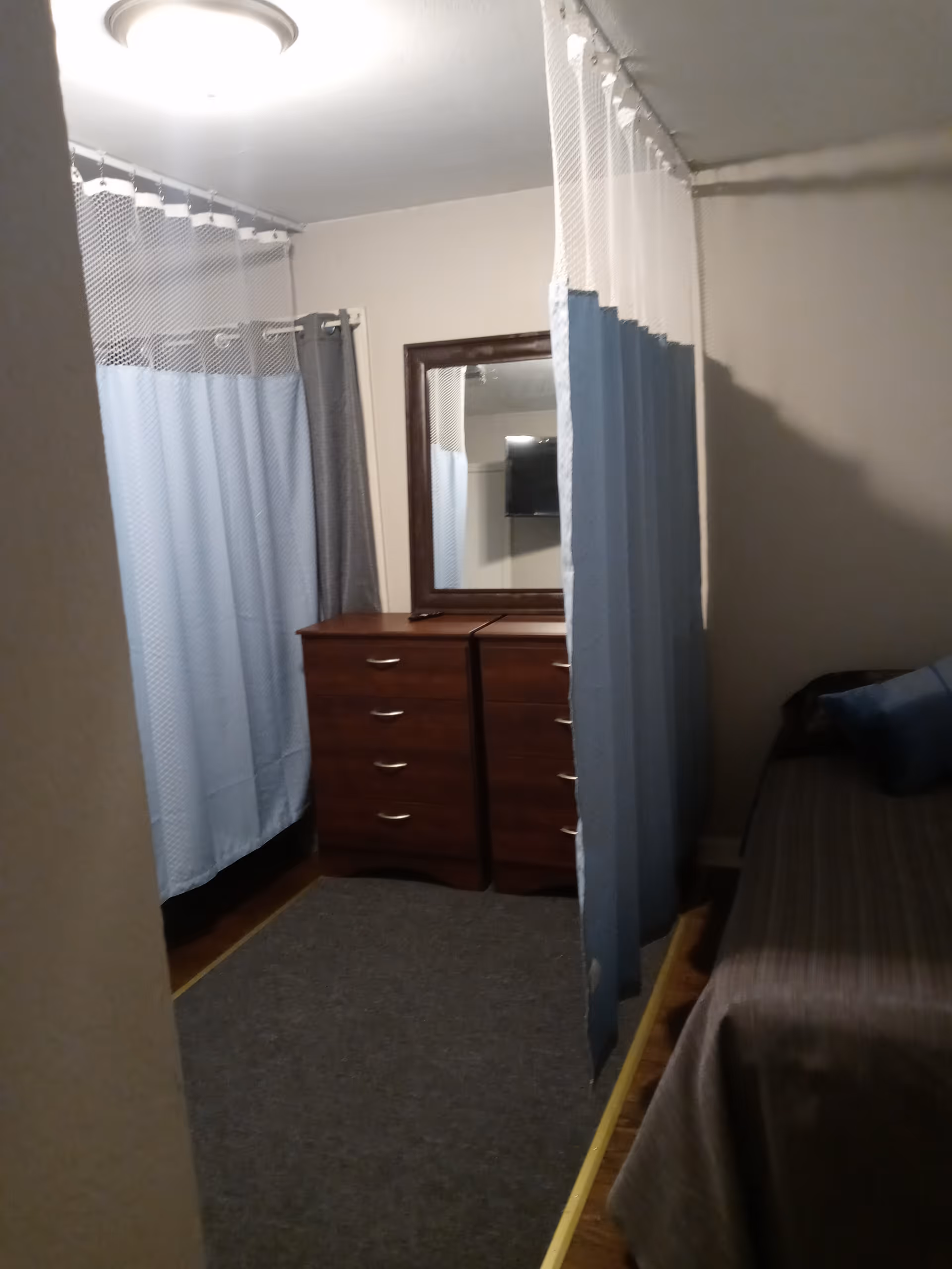 A small room with a bed on the right side covered with a brown blanket and a blue pillow. There are two wooden dressers with drawers against the far wall, topped with a large mirror. Blue and white privacy curtains hang from the ceiling, partially enclosing the space. The room has beige walls and a ceiling light fixture.