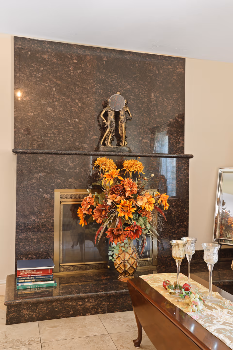 A living room fireplace with a dark brown marble surround and hearth. On the hearth, there is a stack of books on the left side. Above the fireplace, there is a decorative statue of two figures. In front of the fireplace, there is a large vase with orange and red artificial flowers. To the right, a wooden table with a decorative runner holds three ornate glass goblets and some artificial fruit. A mirror is partially visible on the right side of the image.