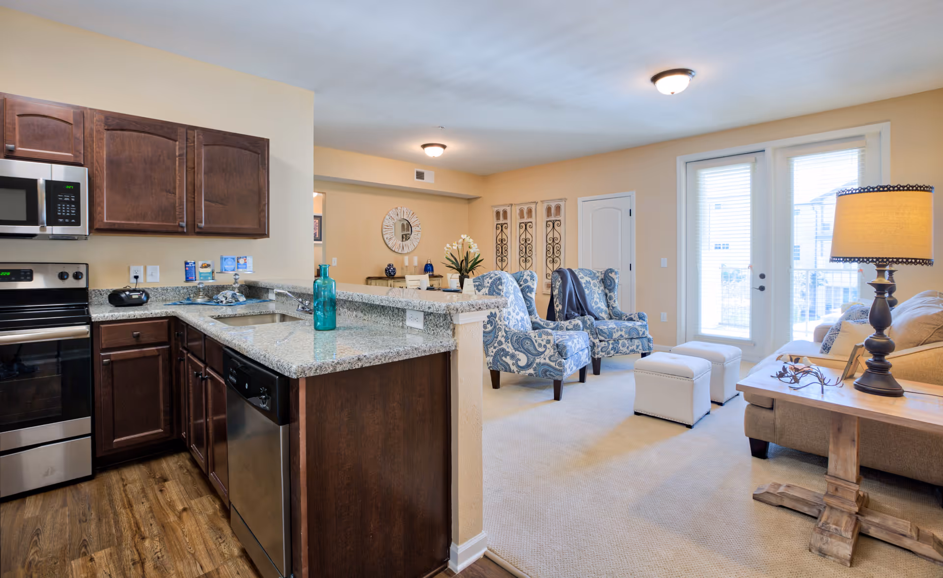 Open-concept living space featuring a kitchen with dark wood cabinets, granite countertops, stainless steel appliances, and a living room area with two blue patterned armchairs, a beige sofa, two white ottomans, a wooden side table with a lamp, and large windows with blinds letting in natural light.