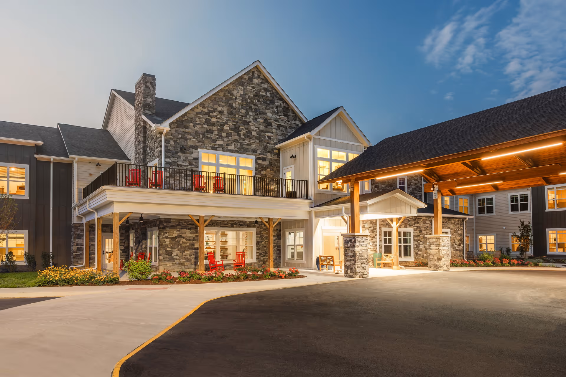 Front entrance of a stone-clad senior living building with an illuminated porte-cochere, balconies with red chairs, and landscaped flowerbeds at dusk.