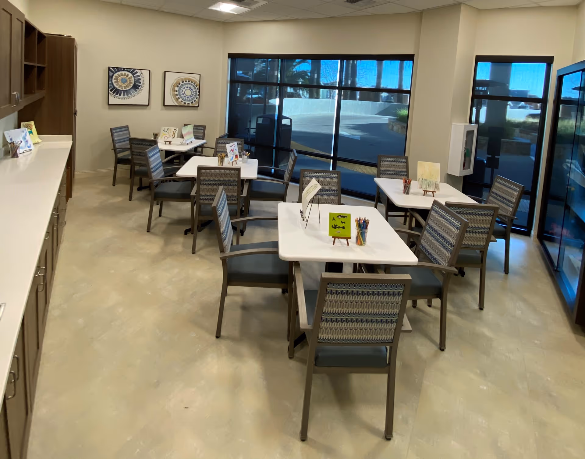 A bright room with several tables and chairs arranged for group activities or dining. Each table has small easels displaying artwork and containers with colored pencils. Large windows let in natural light, and there are cabinets along one wall with additional art supplies or materials.