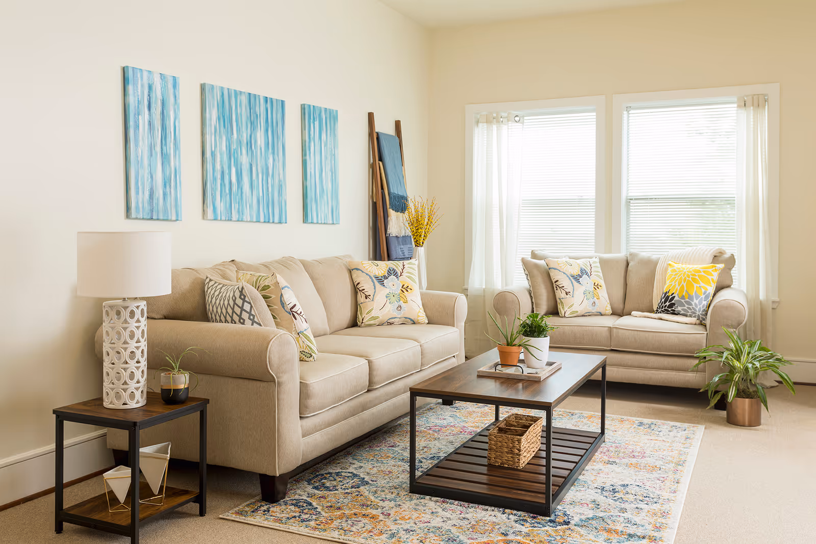 A bright living room with two beige sofas adorned with colorful floral and geometric patterned pillows. A wooden coffee table with a metal frame holds two small potted plants and a wicker basket underneath. A side table with a white decorative lamp and a small plant is next to one sofa. Three blue abstract paintings hang on the wall above the sofa. Two large windows with white blinds and sheer curtains let in natural light. A colorful patterned rug covers the floor, and a tall ladder-style blanket rack with folded blankets and a vase with yellow flowers is in the corner.