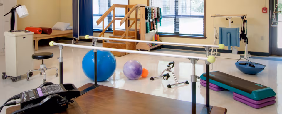 Physical therapy room with parallel bars, exercise balls, a step platform, a small exercise bike, and a wooden staircase with handrails. There is a treatment bed and a stool in the background near a window.