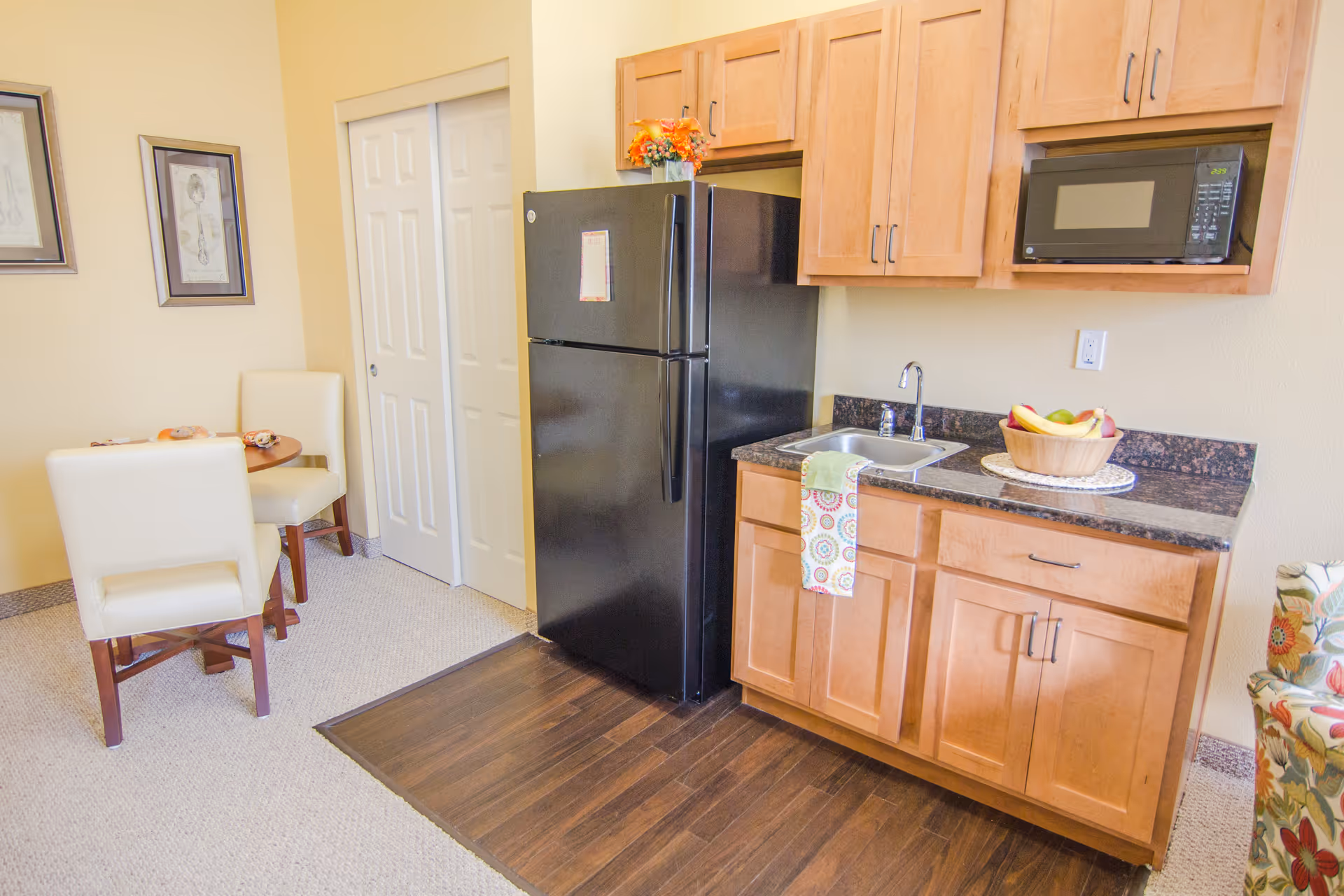 A small kitchen area with light wood cabinets, a black refrigerator, a microwave, and a sink with a colorful towel hanging from the counter. To the left, there is a small round dining table with two cream-colored chairs and framed artwork on the wall. The floor is a combination of dark wood near the kitchen and light carpet in the dining area.
