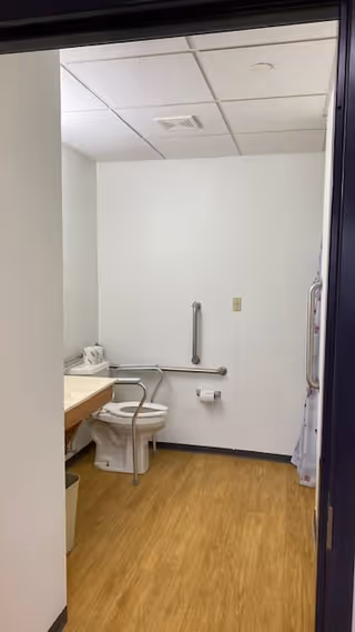 View into a small bathroom with a toilet equipped with a safety rail, a wall-mounted grab bar, a toilet paper holder, a countertop with a tissue box, and a shower curtain visible on the right side. The floor is wood-patterned, and the walls and ceiling are white.