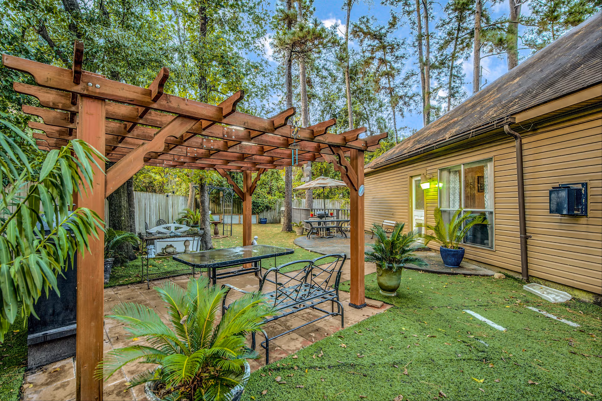 Outdoor patio area with a wooden pergola over a glass table and metal chairs, surrounded by potted plants and greenery. In the background, there is a beige building with windows and a door, and additional seating with an umbrella. Tall trees and a wooden fence enclose the space.