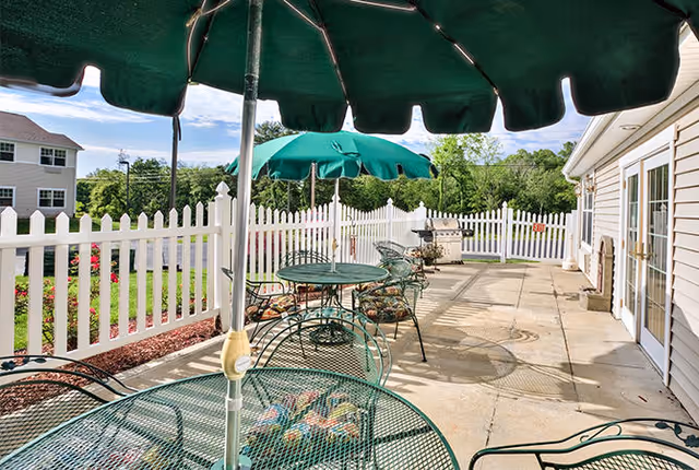 Outdoor patio area with green metal tables and chairs, green umbrellas providing shade, a white picket fence surrounding the space, and a barbecue grill in the background. The patio is adjacent to a beige building with large windows and doors.