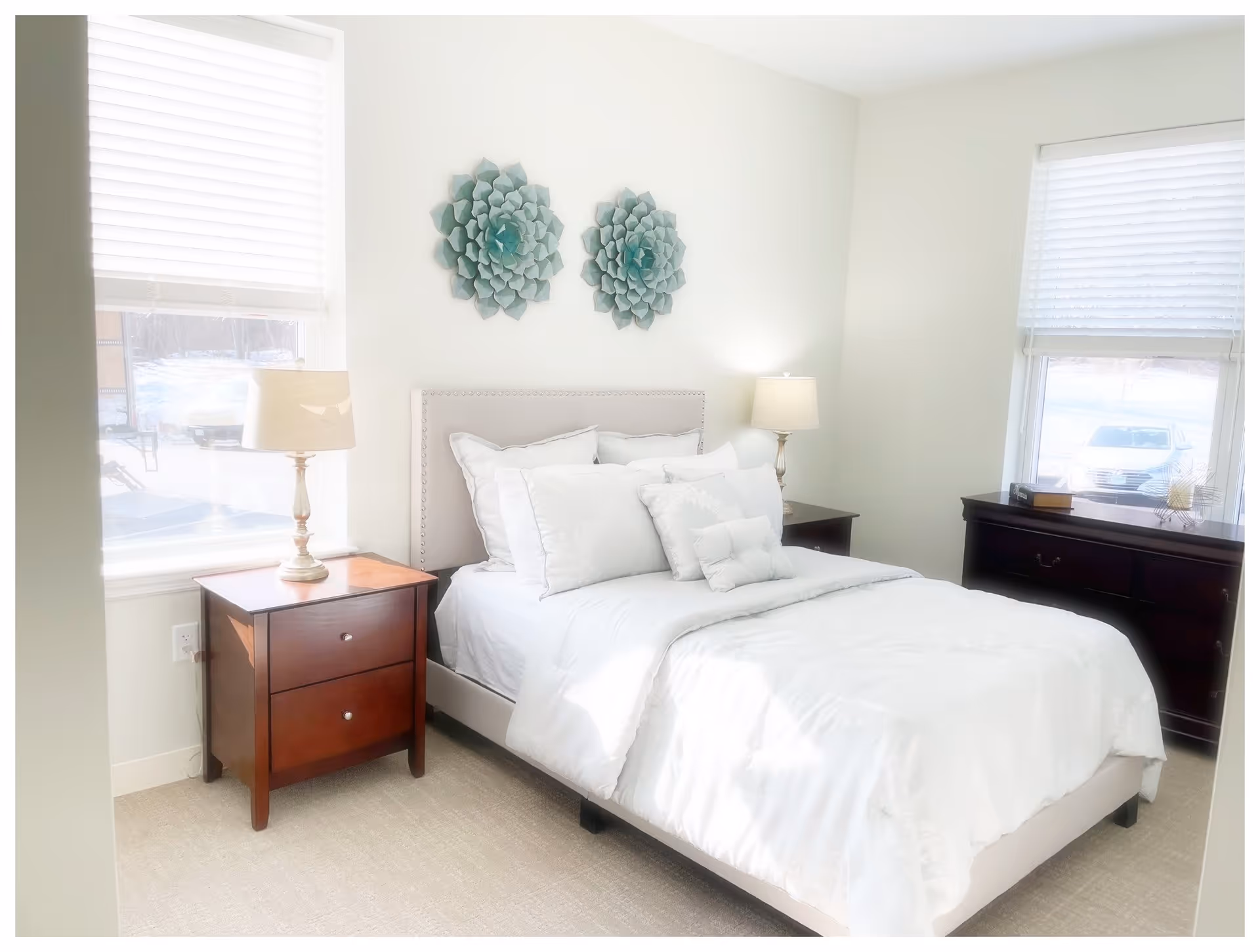 A bright bedroom with a neatly made bed featuring white bedding and multiple pillows. There are two windows with white blinds, one on each side of the bed. On the left side of the bed is a wooden nightstand with a lamp, and on the right side is a dark wooden dresser with some decorative items on top. Two teal-colored floral wall decorations hang above the bed.