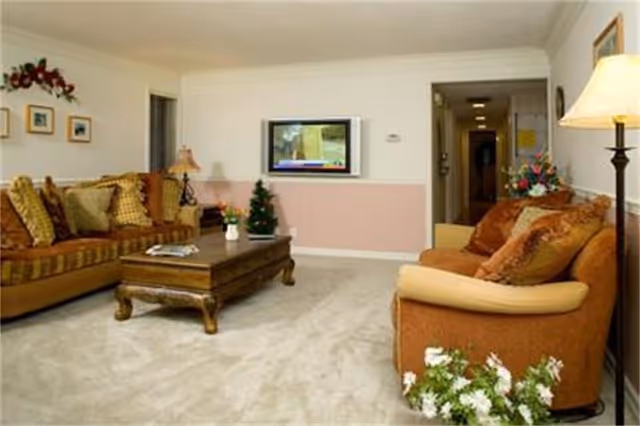 Cozy living room with two upholstered sofas, a wooden coffee table, a wall-mounted TV, lamps, and decorative plants.