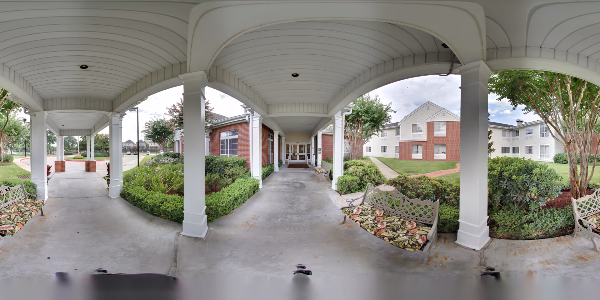 Covered outdoor walkway with white pillars and ceiling, surrounded by green bushes and trees. There are decorative benches with floral cushions on either side of the walkway. In the background, there are multi-story residential buildings with brick and white siding.