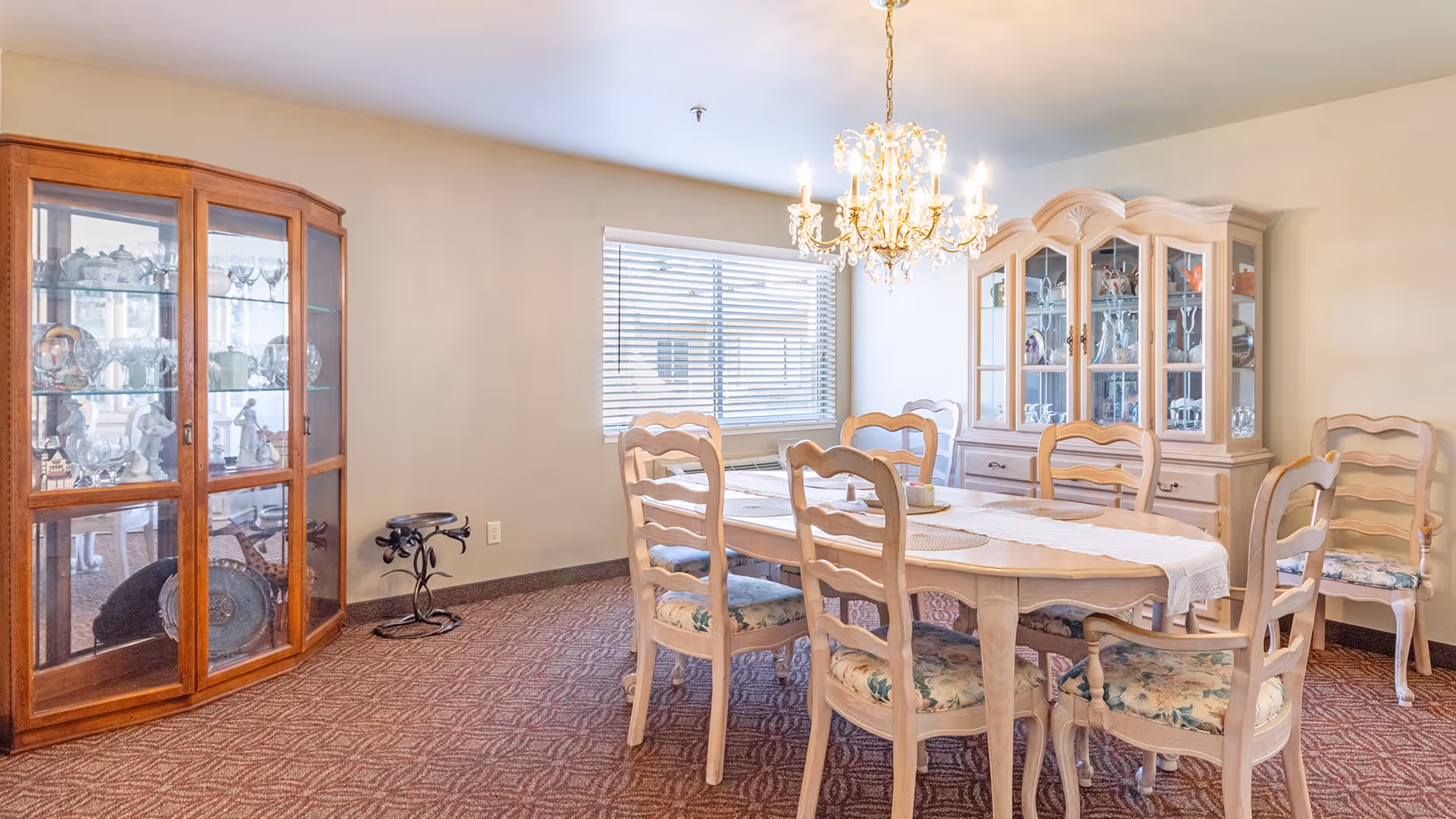 A dining room with a wooden dining table surrounded by six chairs with floral cushions. There is a chandelier hanging above the table. Two glass-fronted wooden cabinets display various glassware and decorative items. A window with blinds lets in natural light, and the floor is carpeted with a patterned design.