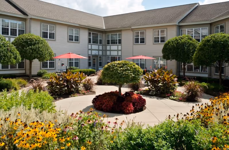 A landscaped outdoor courtyard at a senior living facility with a circular paved walkway, trimmed bushes, colorful flowers, and two red patio umbrellas near the building entrance.