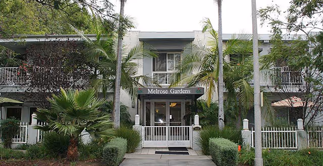 Front exterior view of Melrose Gardens facility with a white fence, palm trees, and greenery surrounding the entrance.