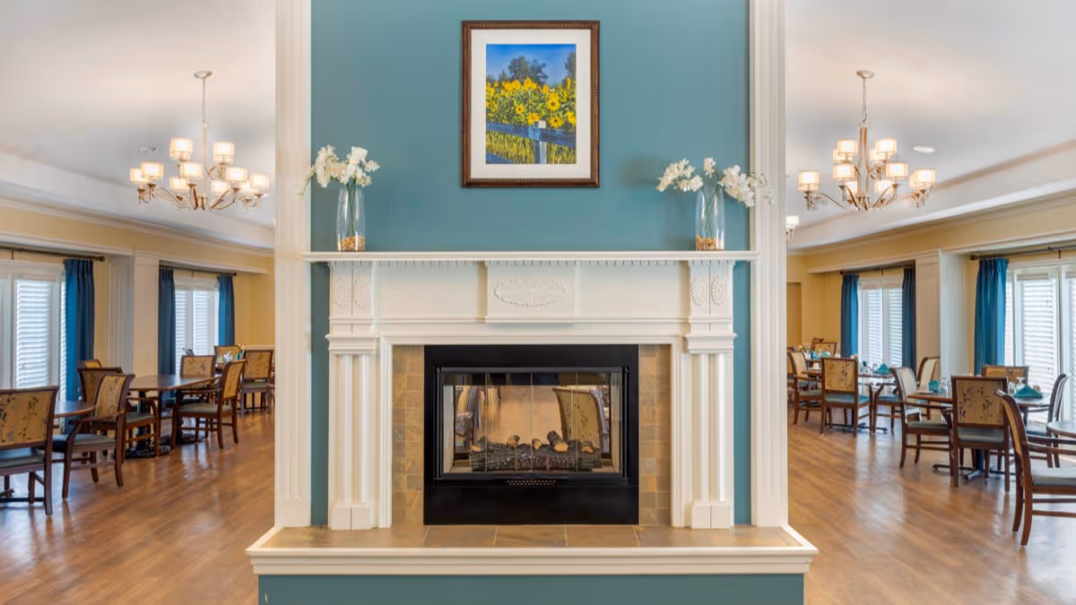 A cozy dining area with wooden floors and multiple tables and chairs arranged neatly. In the center foreground, there is a white fireplace with a black insert, set against a teal accent wall. Above the fireplace hangs a framed picture of sunflowers. Two glass vases with white flowers are placed symmetrically on the mantel. The room is well-lit with chandeliers and natural light coming through windows with blue curtains.