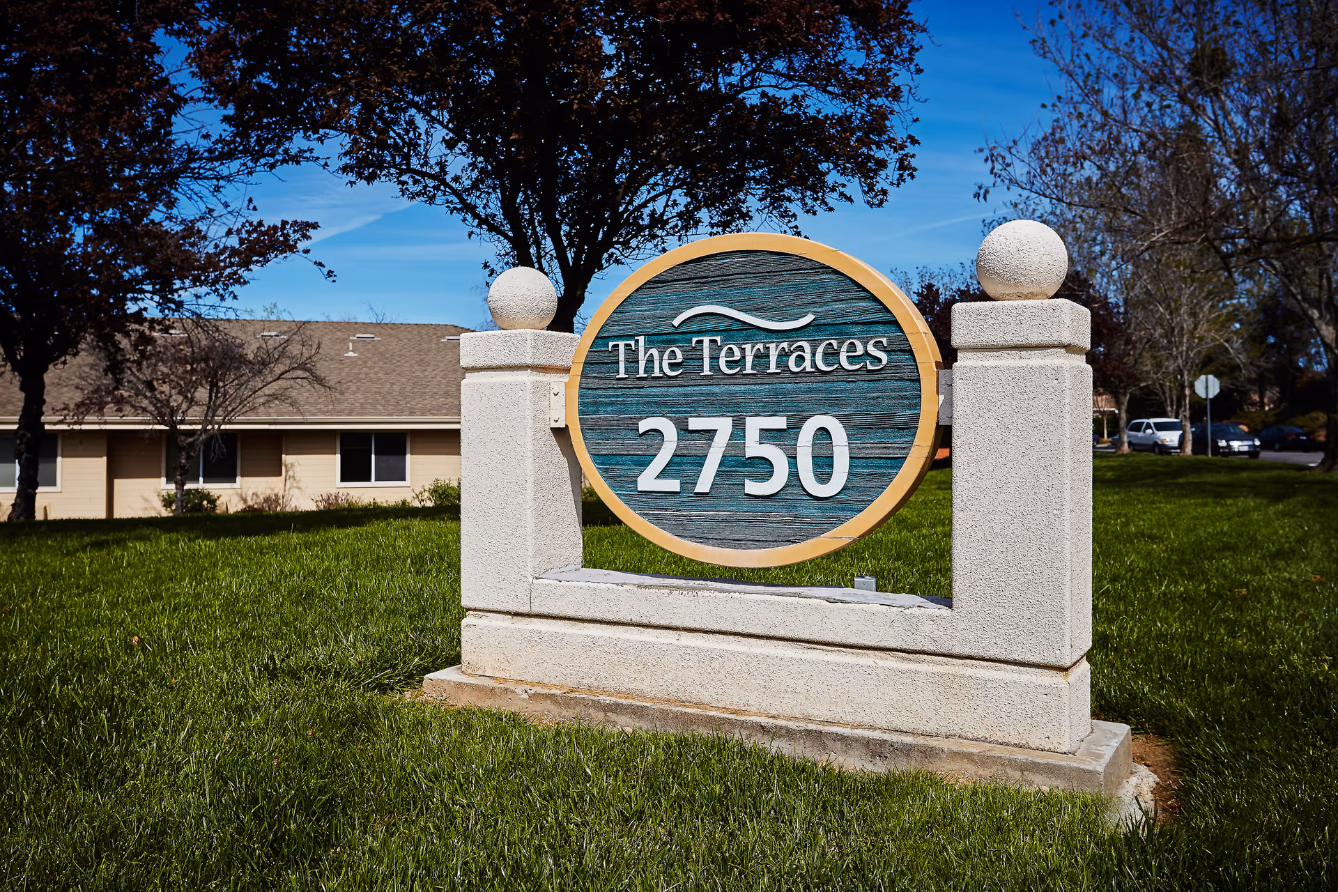 A stone sign on a grassy area with the text 'The Terraces 2750' in white letters on a blue wooden background, with trees and a building in the background under a clear blue sky.