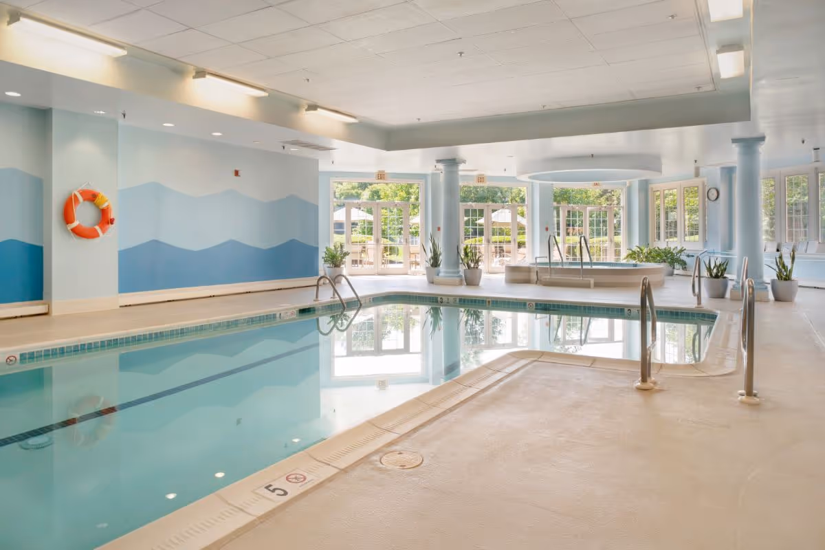 Indoor swimming pool area with light blue walls featuring a wave pattern, a life preserver mounted on the wall, several potted plants, large windows and glass doors letting in natural light, and a hot tub in the background.