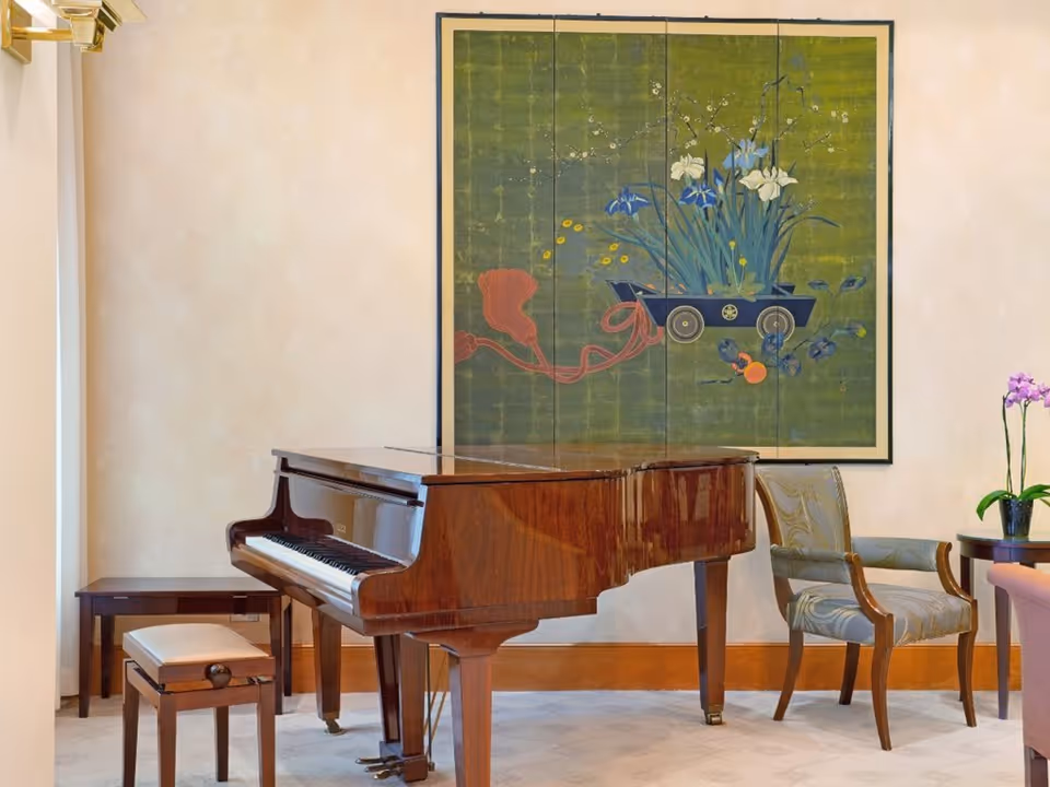 A polished grand piano in a sitting area with upholstered chairs, side tables and a large framed floral painting on the wall.