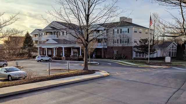Front exterior of a multi-story retirement community building with a covered entrance, parked cars, and an American flag.