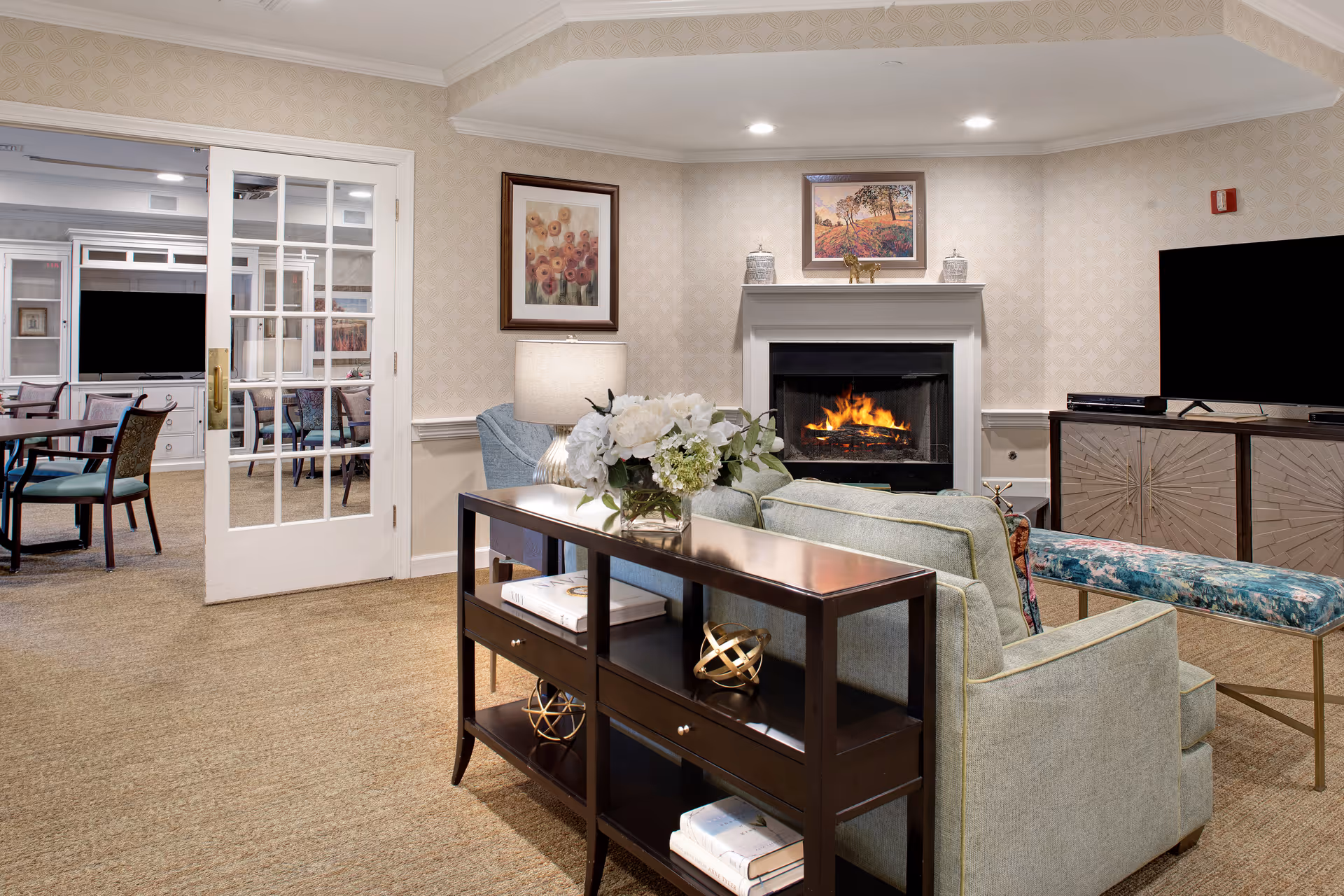 A cozy living room area with a lit fireplace, a large flat-screen TV on a cabinet, a light green sofa, a floral bench, and a dark wooden console table with decorative items and books. There is a lamp and framed artwork on the walls, and a glass-paneled door leading to a dining area with tables and chairs.