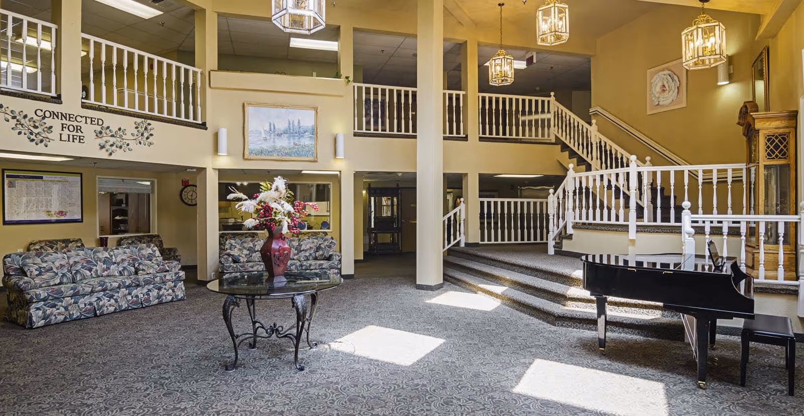 Spacious senior living facility common area with floral patterned sofas, a glass table with a red vase of flowers, a black grand piano, carpeted floor, white railings on stairs and balcony, and warm yellow walls with framed artwork and hanging light fixtures.