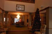 Interior view of a living room area in an assisted living facility with a decorated Christmas tree, upholstered chairs, a chandelier, and a dining table in the background under a high ceiling.