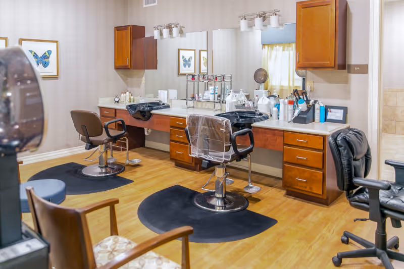 Interior view of a senior living facility spa area with salon chairs, hair washing sinks, wooden cabinets, large mirrors, and various hair care products on the countertops. The room has wooden flooring and framed butterfly artwork on the walls.
