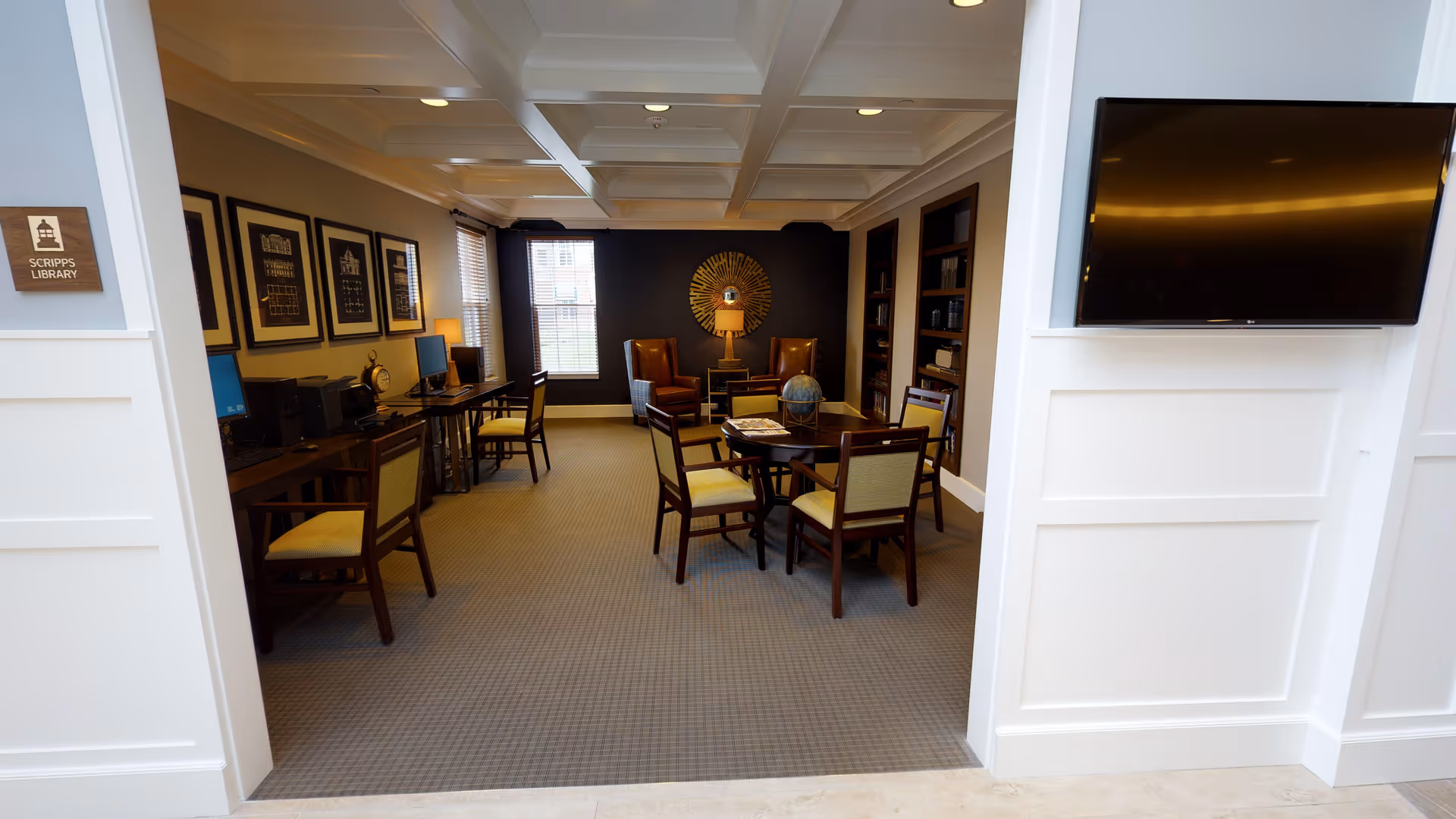 Interior common room/library with a round table and chairs in the center, computer workstations along the left wall, armchairs at the back, and a wall-mounted TV on the right.