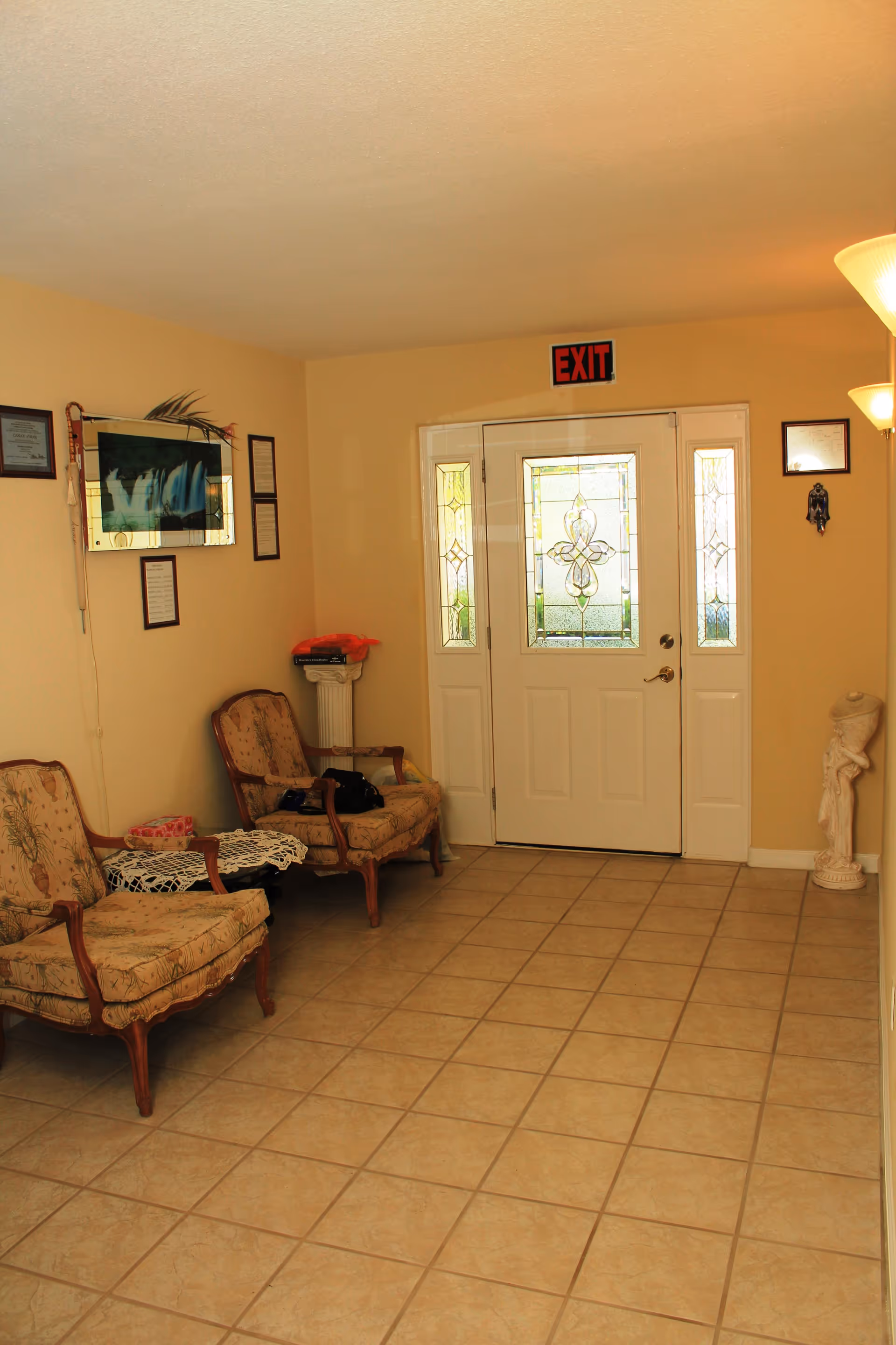 Interior view of a waiting area with two upholstered armchairs, a small table with a lace cover, a decorative column with items on top, and a white door with stained glass panels and an exit sign above it. The walls are beige and there are framed certificates and a mirror hanging on the walls.