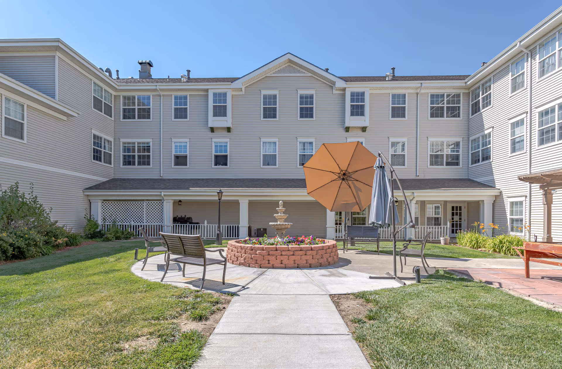 Outdoor courtyard area of a senior living facility with a circular brick flower bed and fountain in the center, surrounded by benches and patio umbrellas. The courtyard is bordered by a three-story building with multiple windows and a covered porch area. The sky is clear and blue.