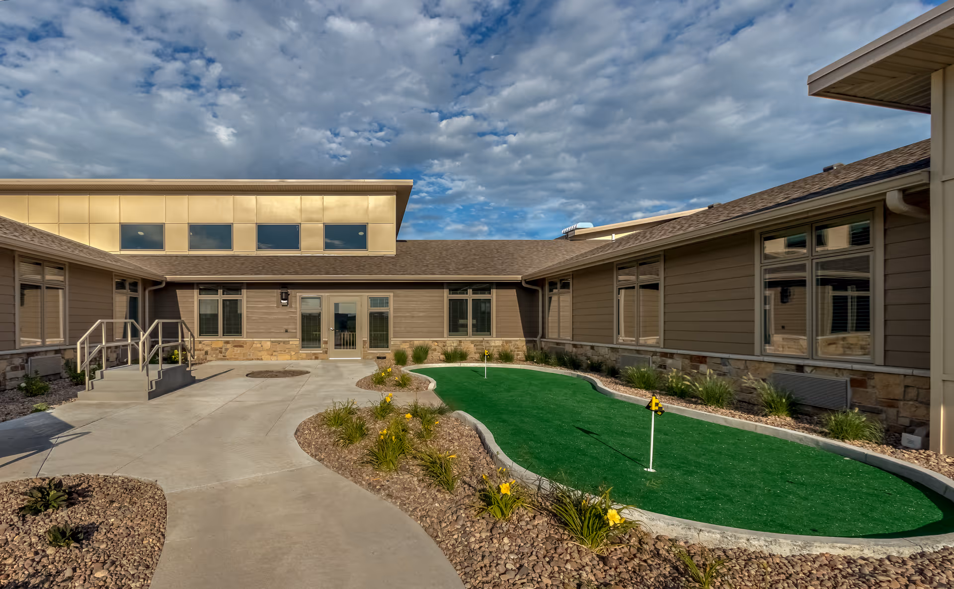 Outdoor courtyard with a small artificial putting green, surrounding walkways and low-rise building wings under a partly cloudy sky.