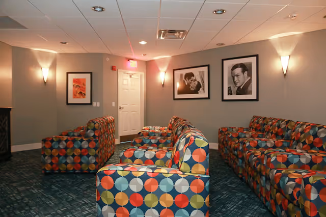 A cozy lounge area with multiple colorful patterned armchairs arranged in rows facing each other. The walls are painted gray and decorated with three black-and-white framed photographs of people. The room is softly lit by wall sconces and ceiling lights, with a white door and an exit sign visible at the back.
