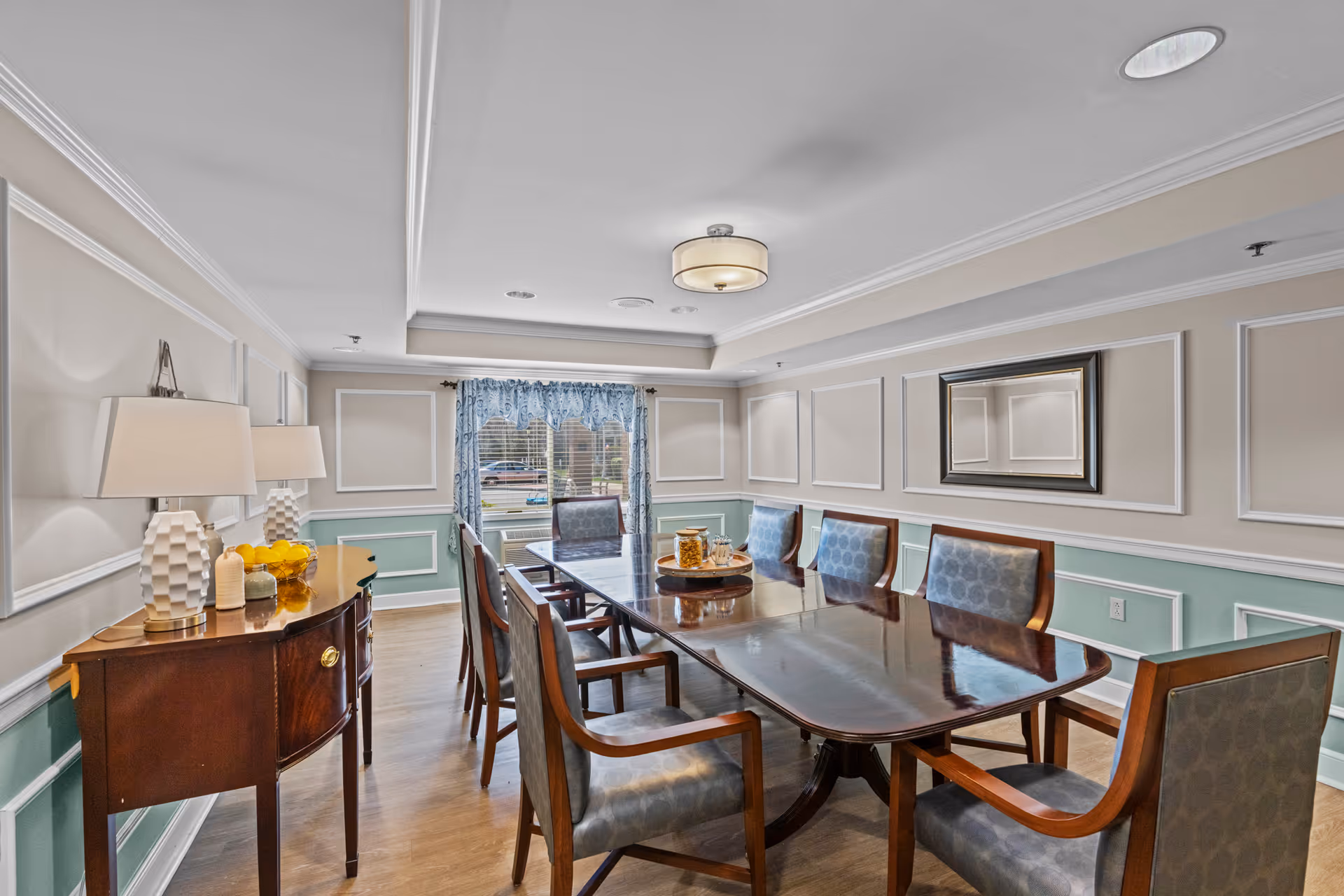 A formal dining room with a long polished wooden table surrounded by eight upholstered chairs. The walls are decorated with white panel molding and painted in beige and light green. A sideboard with two white lamps and decorative items is on the left side. A window with blue patterned curtains is at the far end, and a ceiling light fixture is centered above the table.